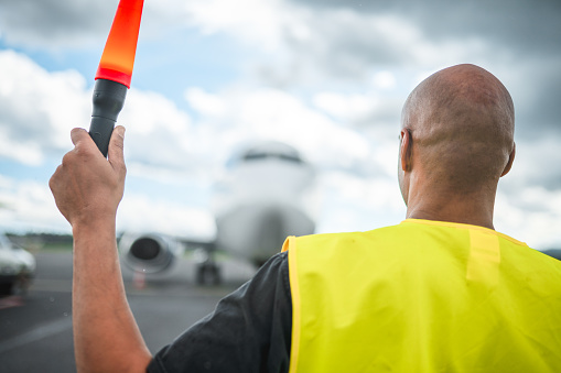 Mixed race airport ground crew member marshalling an aircraft. Wearing a reflective vest and  marshalling with signal lights.
