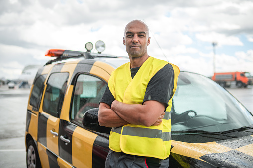 Mixed race airfield operations officer wearing reflective clothes and standing next to an airfield operations car with hands crossed.