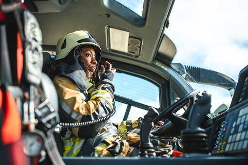 Side view of a Black female airport firefighter sitting in a fire truck using a CB station while driving towards an emergency location at the airport runway.