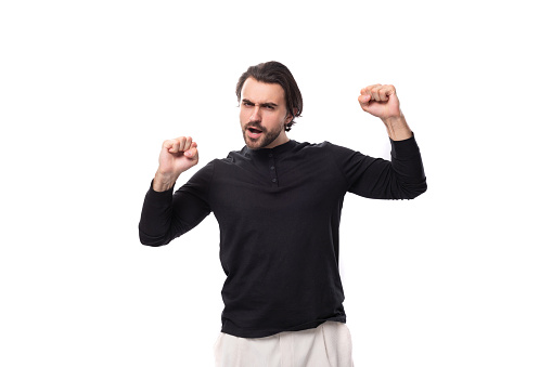 portrait of a dancing positive young handsome brunette man with a hairstyle and beard dressed in a black jacket isolated on white background with copy space.