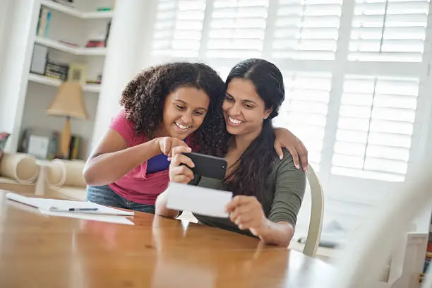 Happy teenage daughter and mother depositing check through smart phone at home