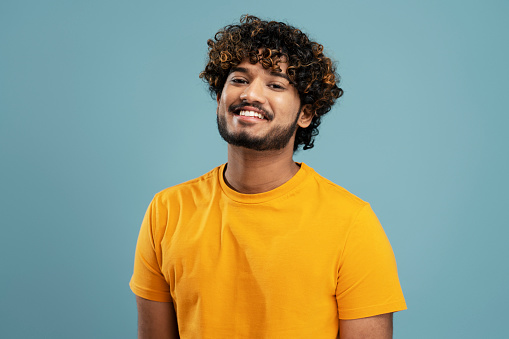 Successful confident easy-going curly haired bearded young Hindu man, wearing a bright yellow t-shirt, smiling a beautiful toothy smile looking at camera, isolated over blue background