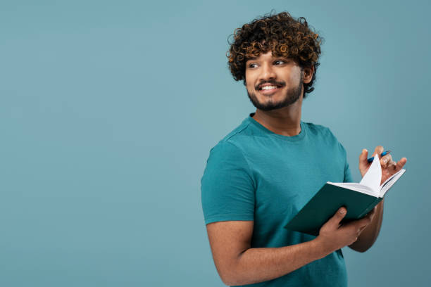 istock Indian male writer writing in notebook looking away isolated on blue background. Education concept 1699441371