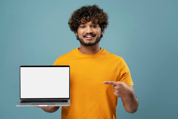 istock Smiling indian man using laptop pointing finger on white display isolated on blue background 1699441377
