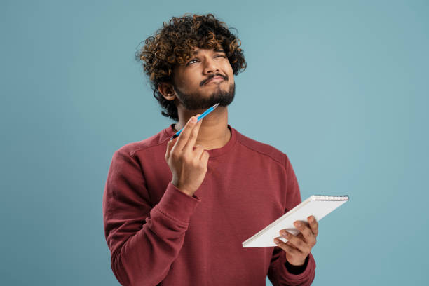 istock Pensive Indian man dreamily looking up posing with notepad in his hand, isolated on blue background 1699441942