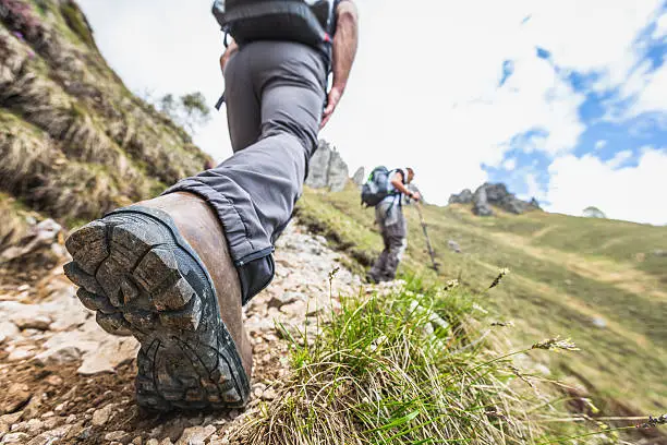 Hikers walk on a mountain trail.  The focus of the image is on one hiker's right boot.  The sole of the boot is in the foreground of the image.  The hiker wears jeans and a backpack.  Another hiker is visible in the background, walking on the dirt trail.