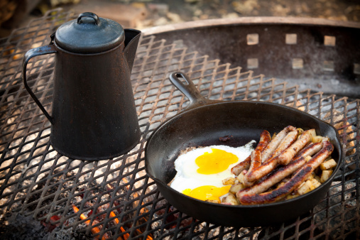Coffee, eggs and sausage breakfast cooking over a campfire. Cast iron fry pan and coffee pot are on a steel grate placed directly over the flame and coals. No people in photo. High resolution color photograph. Horizontal composition.