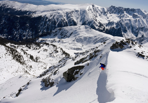 Free skier beginning his descent with a big drop off a cornice. His jump is captured on a beautiful panorama background with snow covered peaks. The weather is quiet, the sky is clear and the snow is just perfect for free ride skiing.