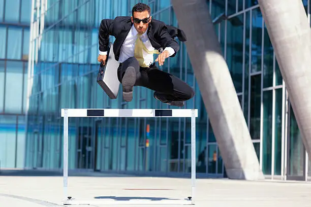 young business man with briefcase and sunshades running over hurdle
