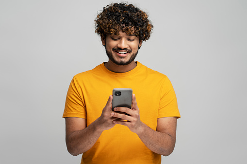 Asian hipster with mobile, chatting, communication, ordering food isolated on gray background. Young smiling Indian man holding smartphone using mobile app shopping online. Mobile banking concept