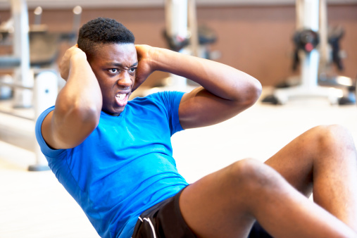 A young coloured athlete, doing sit ups in a fitness studio,with a strained facial expression.