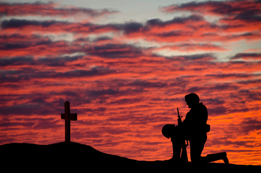 A soldier kneeling in front of a grave with a cross at sunset.