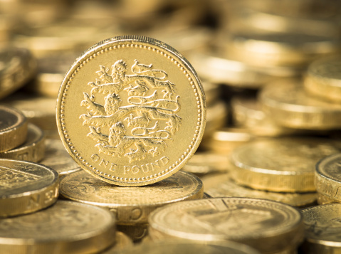 Focus on one British Pound coin, featuring three heraldic lions, among a pile of pound coins.