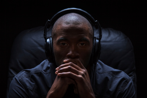 Dramatic photograph of a black man sitting in a leather chair listening to music with over-ear headphones.