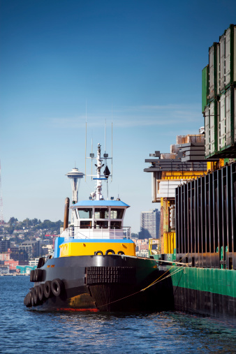 A brightly colored tug boat anchored to a large container-carrying barge in the Seattle Sound with the Space Needle in the background