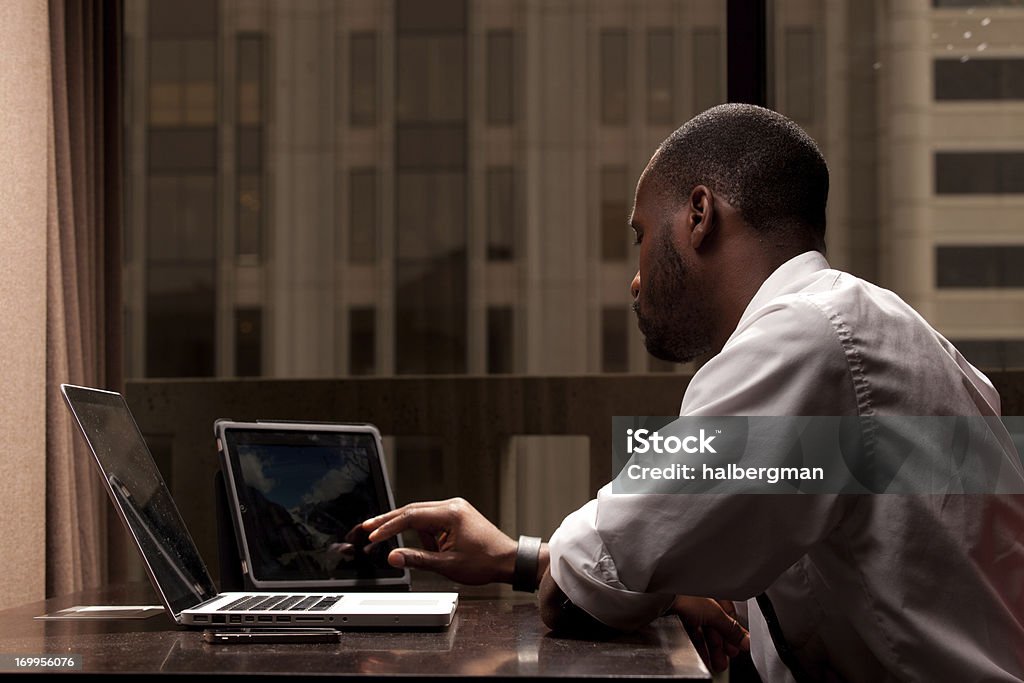 Man Working on a Tablet PC A black businessman surrounded by tech gadgets working on a tablet computer from his hotel room African Ethnicity Stock Photo