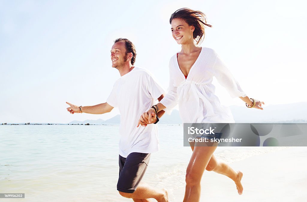 Feliz pareja sosteniendo las manos y disfrutar de corriendo en la playa. - Foto de stock de Azul libre de derechos