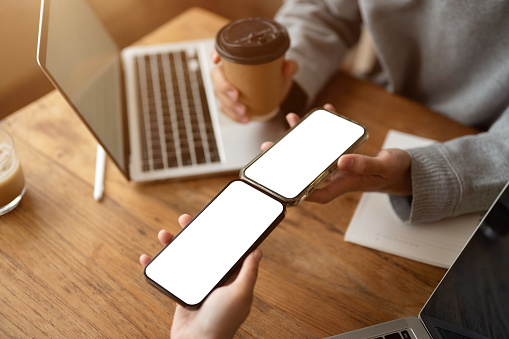 Close-up image of a woman is sharing files or a contact on her smartphone to her friend's smartphone while sitting in a coffee shop together. Smartphone white screen mockup. Technology concept