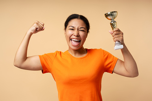 Young overjoyed African American woman holding trophy cup, celebration success isolated on beige background. Sport victory, winning competition concept