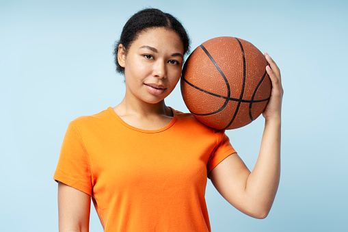 Portrait of beautiful smiling African American woman playing basketball, holding ball looking at camera, isolated on blue background. Sport, hobby, healthy lifestyle concept