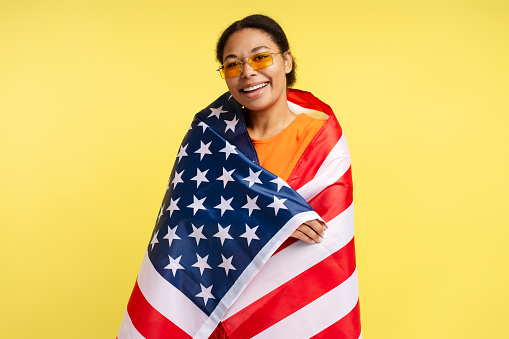Portrait of confident smiling African American woman holding American flag isolated on yellow background, celebration Independence day. July 4th concept