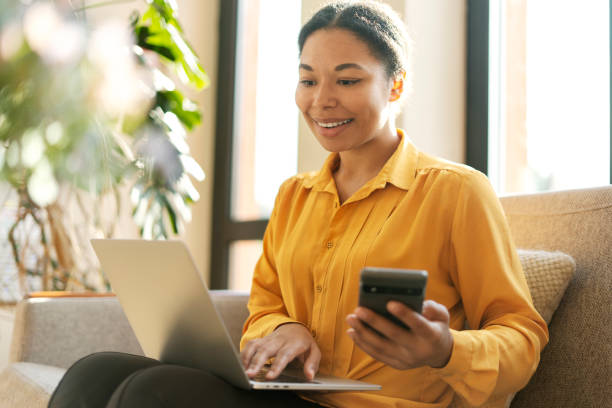 istock Smiling African American woman, freelancer, manager holding mobile phone, using laptop working online at modern workplace. Female online shopping, ordering food sitting at home. Technology 1699593266