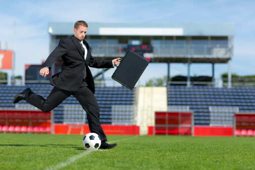 soccer player in business suit with suitcase kicking ball on football playing field