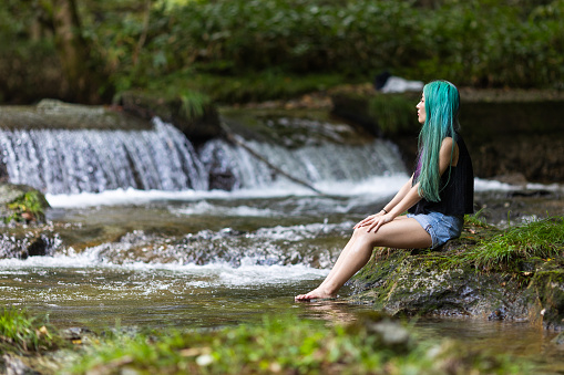 A young Japanese woman with long straight hair dyed in two colors enjoying nature by a river and waterfall with feet in the water.