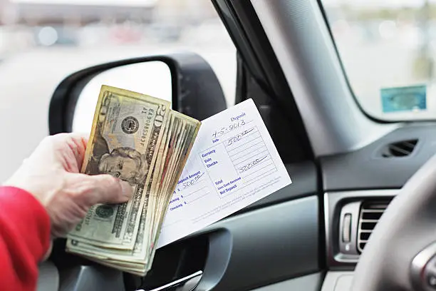 A man in his car is holding five hundred dollars in US twenties and a bank deposit slip while waiting in line for a bank drive through teller window. Could represent a small business transaction, or personal home finances, etc.