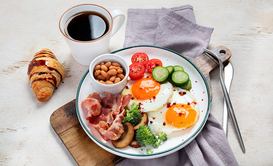 Traditional Englis breakfast plate with bacon strips, sunny side up eggs, vegetables and cake on light background.