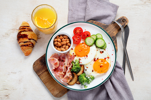 Traditional Englis breakfast plate with bacon strips, sunny side up eggs, vegetables and cake on light background.