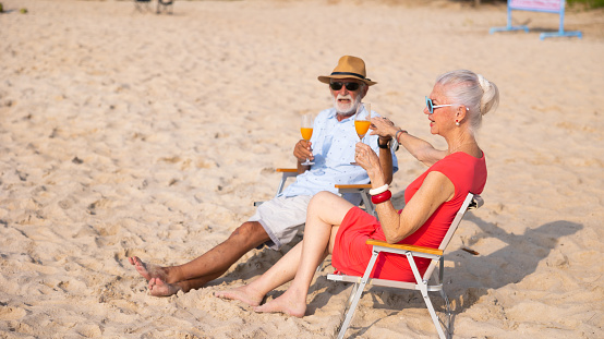Elderly couple man and woman wearing fashion sunglasses talking together and looking at the sea sky sitting on chair on beach. Vacation trip summer holiday.