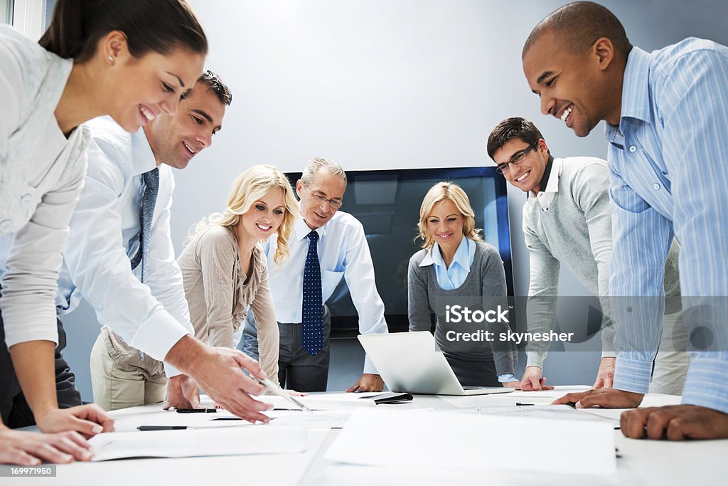 People standing around a table with a laptop A large cheerful group of successful businesspeople standing on a meeting in the office and looking at reports.     Community Stock Photo
