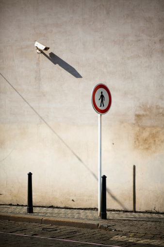 A strange juxtaposition of an outdoor security camera seemingly pointed at a European-style round pedestrian crossing sign with the icon of a walking man.