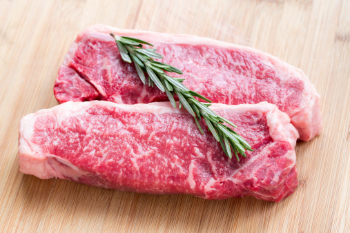 Two raw Kobe beef New York steaks on a bamboo cutting board with a sprig of Rosemary