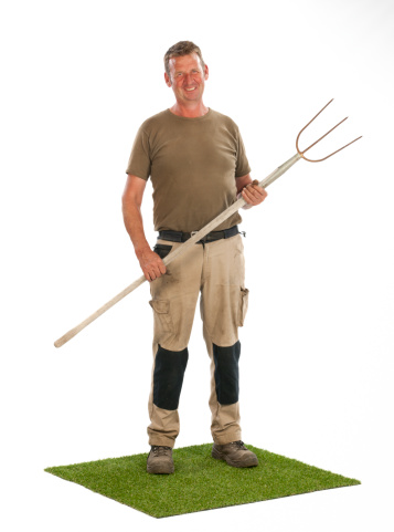 full length shot of a smiling farmer holding a pitchfork standing on grass looking at the camera on white background