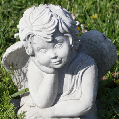 A cheerful, chubby child angel (putto) gazes patiently in the grass of a cemetery in Merton, Surrey, England. A statue of this kind will overlook the grave of a child. This image is in exact square proportion.