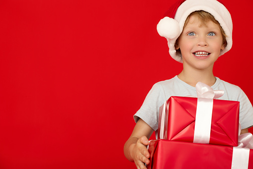Funny kid with a smile on his face in a red hat Santa holds Christmas gift in his hand. Happy Baby in Santa Red Hat Keeps Christmas Presents