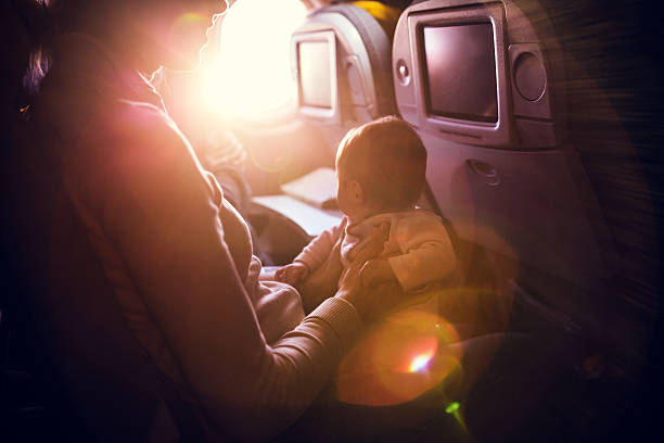 Airplane Travel With Infant A mother and her baby girl sit in a passenger airline seat, the sun shining brightly in through the plane window.  While air travel with children can be difficult, both mom and child are content, the mother with a smile on her face. Horizontal image.  INTENTIONAL LENS FLARE. 2 5 months photos stock pictures, royalty-free photos & images