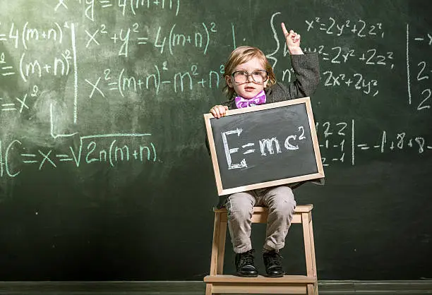 Brainy little boy sitting in front of blackboard. Boy showing the mass-energy equivalence formula on small chalkboard