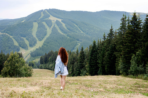 A beautiful young girl stands against the background of the mountains in the summer. Beautiful landscape of mountains. The concept of tourism and travel.