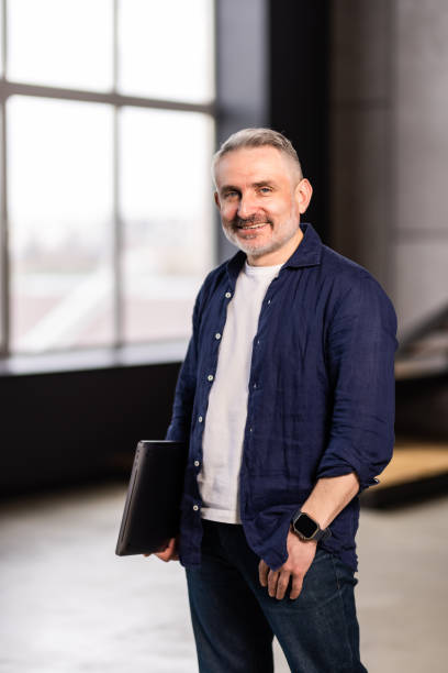 istock Mature man holding using laptop standing leaning on white wall, posing looking at camera. 1699843205