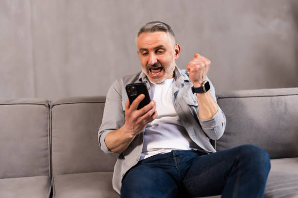 istock Overjoyed laughing middle aged man looking at cellphone screen, reading message with amazing news, celebrating winning online lottery gambling auction, sitting on sofa at home. 1699852634