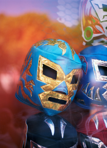 This is a vertical color photograph of an assortment of Lucha Libre wrestling masks in a San Francisco window display. The glass has scratches and reflects trees and sky in the predominantly orange background. 
