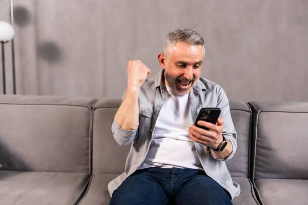 istock Emotional bearded middle-aged man celebrating success, gambling or trading online, using mobile phone while sitting on couch at home. Online gaming, financial markets concept 1699859727