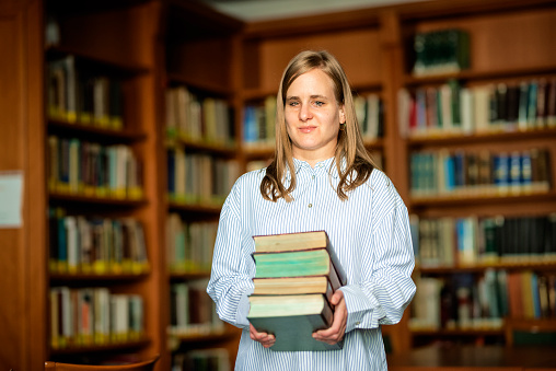 Shot of a blond haired visually impaired woman standing in the library holding books in her hands. She is working in the library.