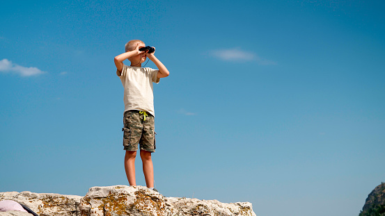 Child boy looks through binoculars at the top of the mountain. A child hiker traveler inspects the surroundings through binoculars while traveling on vacation. Active lifestyle, healthy childhood.