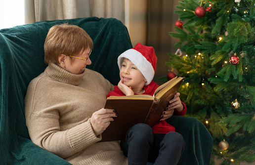 Grandmother and grandson in Santa's hat read fairy tales on the background of the Christmas tree.