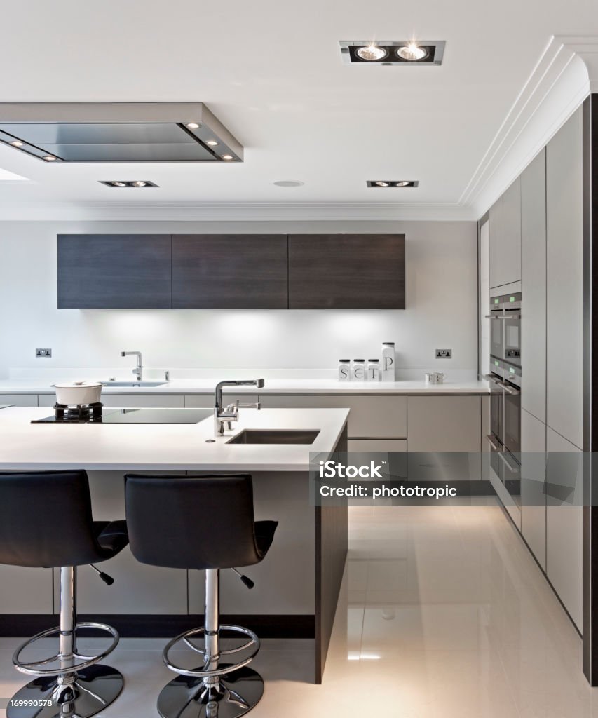 bright modern kitchen a near square composition of a modern kitchen brightly lit by both natural light coming in from the left and internal lighting. A large kitchen island sits in the foreground with two leather chairs. Next to the sink is a tap with filtered water and also a boiling water-on-demand tap. This is a beautifully fitted kitchen with walnut and caramel coloured cupboards and cream coloured marble flooring. Illuminated Stock Photo