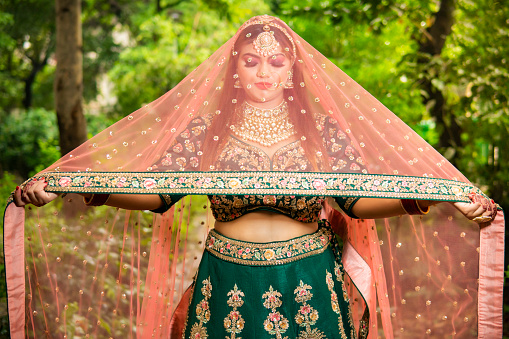 Outdoor portrait of beautiful Indian bridal wearing traditional lehenga, jewelry. She is holding her dupatta above head and standing against beautiful lush green nature.